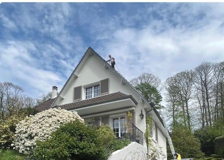 Couvreur debout au faîtage d'une maison à toiture en tuiles, nacelle télescopique déployée sur le côté du bâtiment